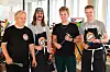 Fire smilende personer poserer sammen i et lyst innendørslokale med T-skjorter og forklær.