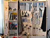 Åpent skap med håndverktøy, arbeidsklær og smådeler i et verksted.
