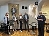 Fem personer står samlet i et lyst galleri med kunstverk og stoler.