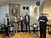Fem personer står samlet i et lyst galleri med kunstverk og stoler.