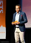 Frode Skaarnes i blå blazer holder papirer på en scene med oransje banner og projisert bakgrunn.